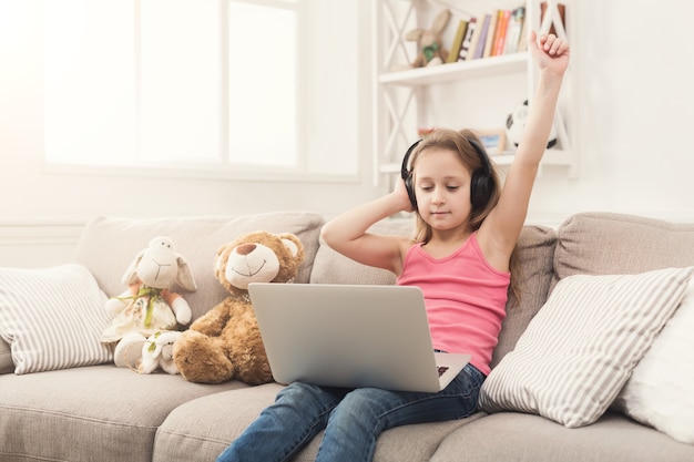 Happy little girl wins in computer game. Cute kid in headphones playing online on laptop, sitting on couch at home with hand raised in the air, copy space