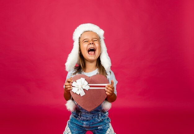 Happy little girl in winter hat with christmas present on pink background