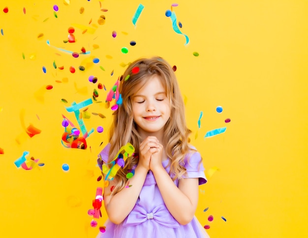 Happy little girl with blond hair and in a blue dress catches confetti on a yellow background, holiday concept