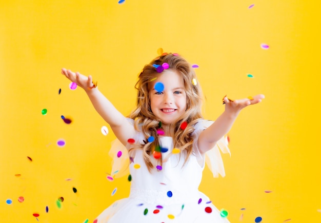 Happy little girl with blond hair and in a white dress catches confetti on a yellow background, holiday concept