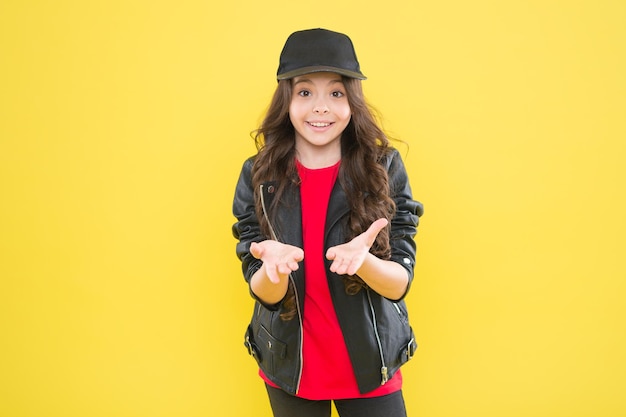 Happy little girl with cool look in fashion wear hold open hands yellow background presenting product