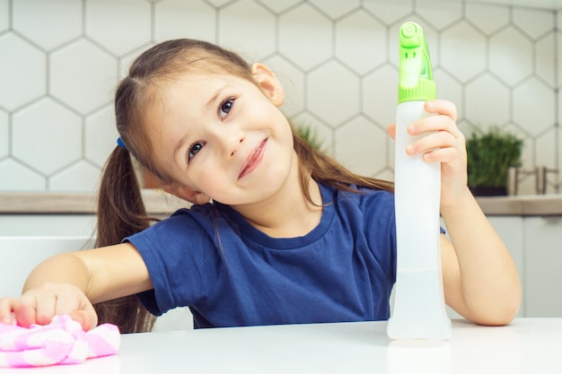 Happy little girl with detergent sprayer and household rag at table Portrait of child tidying up kitchen home cleaning