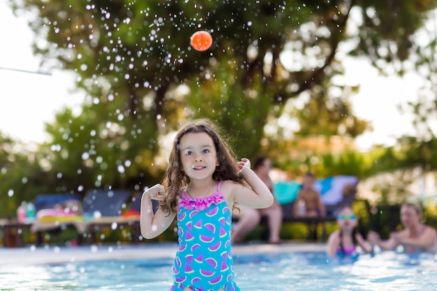 Happy little girl with her hair down in a bright swimsuit playing ball in the pool on a Sunny summer day