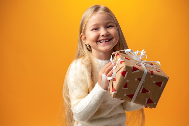 Happy little girl with long hair holding a gift box on a yellow background