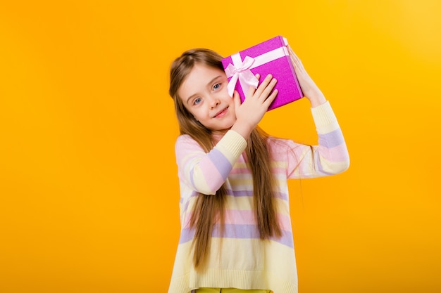 Happy little girl with long hair holding a pink gift box on a yellow space isolate