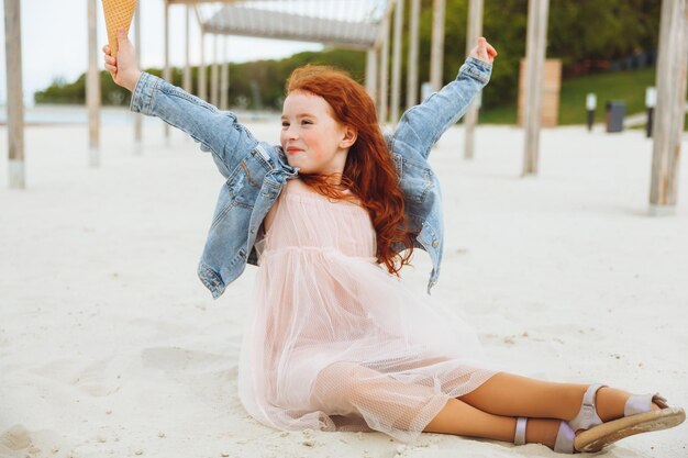 Happy little girl with red hair eats ice cream on the beach in summer summer vacation