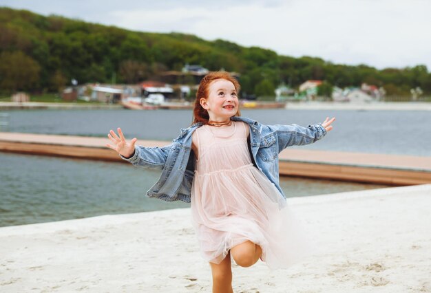 赤い髪の幸せな少女は、ビーチでビーチの子供に沿って走ります