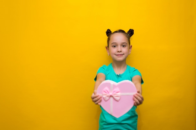 Счастливая маленькая девочка с положением хвостов изолированная над желтой стеной держа ходя по магазинам розовую сумку. задумчиво улыбается