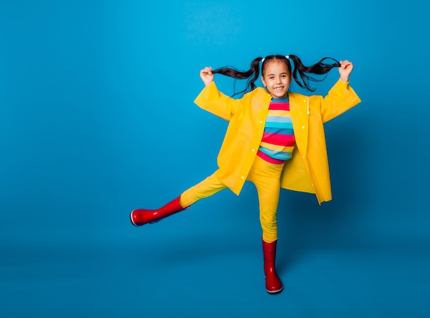 Happy little girl in a yellow raincoat and red boots on a blue background.