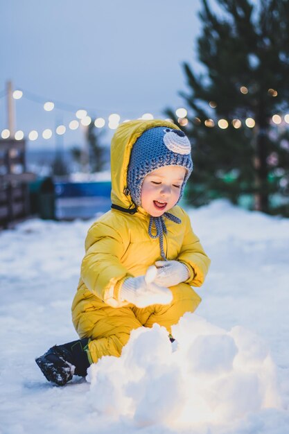 счастливая маленькая девочка в желтом зимнем комбинезоне вязаной шапке весело играет со снегом в зимний день в парке