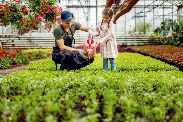 Happy little girl and young florist using watering can and watering plants together in a greenhouse