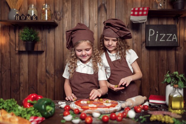 Счастливые маленькие девочки с скалкой и шпателем, улыбаясь и глядя на камеру, готовя вкусную пиццу в уютной кухне вместе