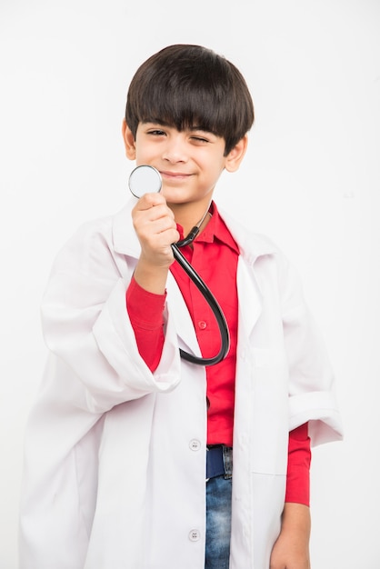 医師として医療制服を着て、聴診器を保持し、白い背景で隔離のカメラを見て幸せな小さなインドのアジアの少年
