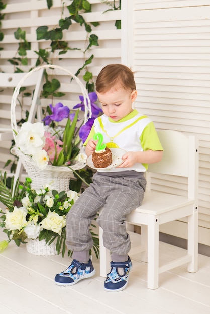 Happy little kid boy celebrating his birthday holds piece of cake, indoor. Birthday party for children. Carefree childhood, happiness. Two years old.