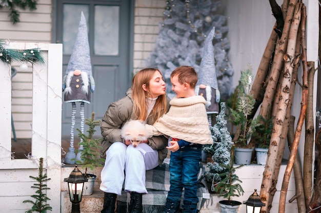 Happy little kids with white little puppy sitting on the porch of the Christmas decorated house, snowing outdoor. Happy New Year and Merry Christmas. Magic winter