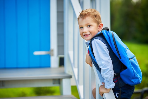 Счастливый маленький мальчик дошкольного возраста с рюкзаком, позирует на открытом воздухе