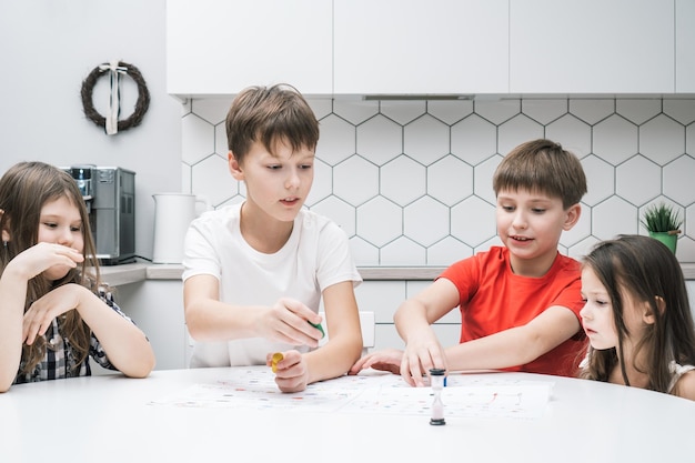 Счастливые маленькие школьные друзья играют в настольную игру на бумажной карте с кубиками и чипсами за кухонным столом Развлекательная настольная игра