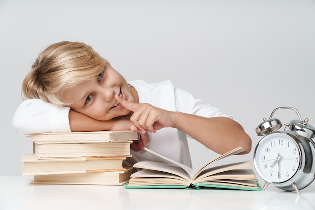 Felice piccolo scolaro che fa i compiti alla scrivania, leggendo libri isolati su un muro grigio