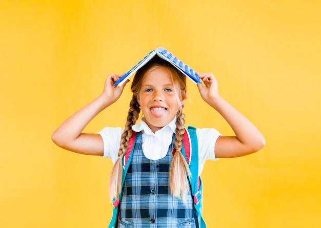 Photo happy little schoolgirl with a book on her head, standing on a yellow background.