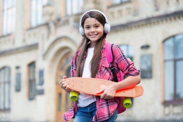 Happy little skater girl hold skating board listening to music in headphones summer urban outdoors, penny.