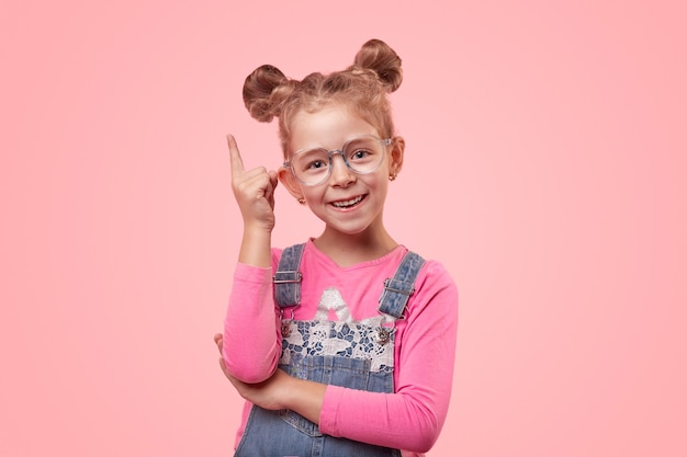 Happy little smart girl in denim overall and spectacles pointing up and looking at camera against pink background