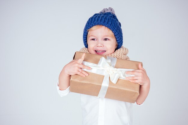 Happy little smiling blonde curly hairstyle boy in a knitted blue hat with christmas gift box with bow on white background in studio. new Year present in the hands of a male child