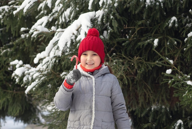 Фото Счастливая улыбающаяся девочка в красной шапке, стоящая рядом с елей и наслаждающаяся конфетной тростью