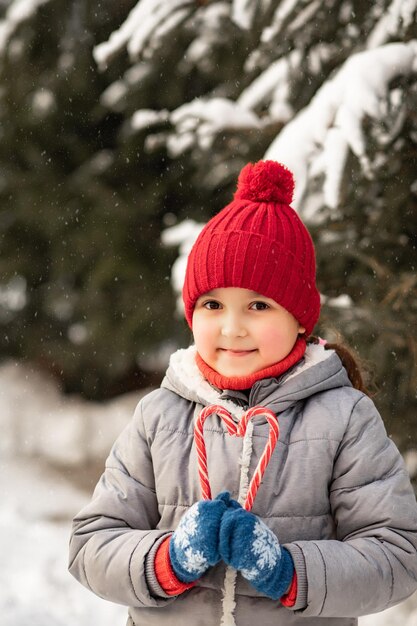 Счастливая улыбающаяся девочка в красной шапке, стоящая рядом с елей и держащая конфеты в форме сердца