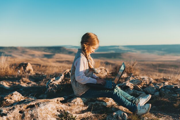 Happy little teen girl sits on a hill with a laptop. Digital nomads
