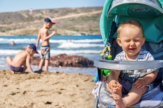 Happy little toddler enjoying sunlight in stroller while brothers playing on seashore in summertime