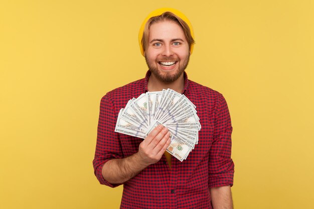 Happy lottery winner rich hipster guy in beanie hat and checkered shirt holding dollars and looking at camera satisfied toothy smile great profit money savings studio shot isolated on yellow