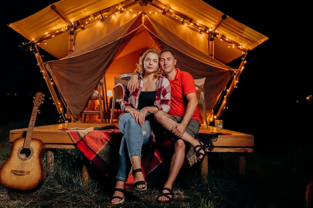 아늑한 모닥불 근처에서 저녁에 글램핑을 하며 휴식을 취하는 행복한 사랑스러운 커플 야외 레크리에이션 및 레크리에이션 라이프스타일 컨셉을 위한 럭셔리 캠핑 텐트