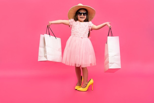 Happy lovely moments of shopping time with cute little girl in dress standing in mother`s big shoes with white packages in hands isolated on pink background.