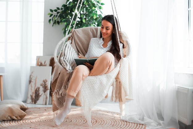 Happy lovely relaxed trendy woman with long healthy hair reads a book in stylish clothes in the modern living room while sitting on hanging chair