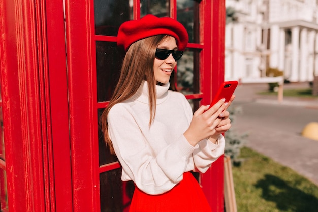 Happy lovely stylish girl with lightbrown straight hair is wearing red beret and white pullover in sunglasses is using smartphone on sunny street