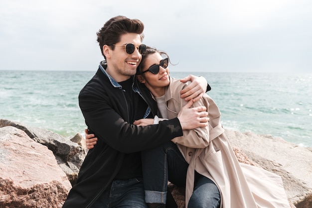 Happy lovely young couple wearing coats sitting at the beach, holding hands, embracing