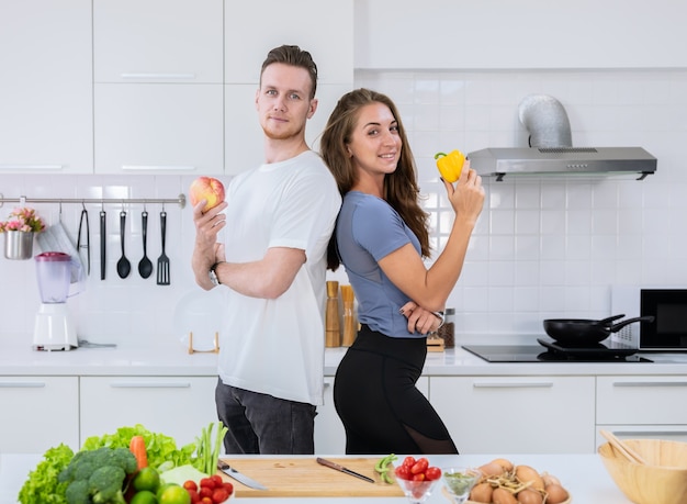 Happy lover couple cooking together in kitchen. Young man and woman standing and holding fresh fruit and vegetable in their hands