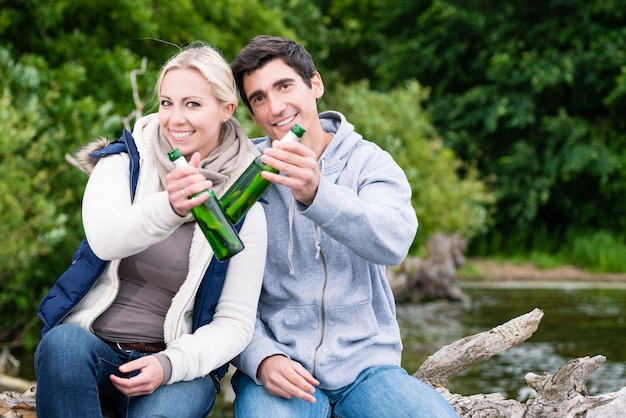 Happy lovers in vacation sitting at waterside on a trunk clinking beer bottles