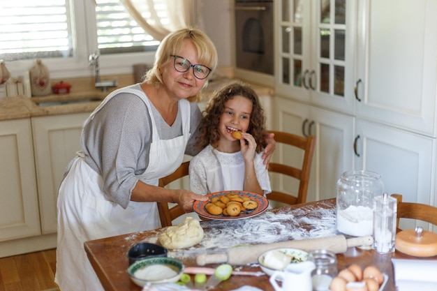 Felice amorevole nonna e sua nipote stanno preparando la panetteria insieme, divertendosi in cucina, guardando la macchina fotografica.