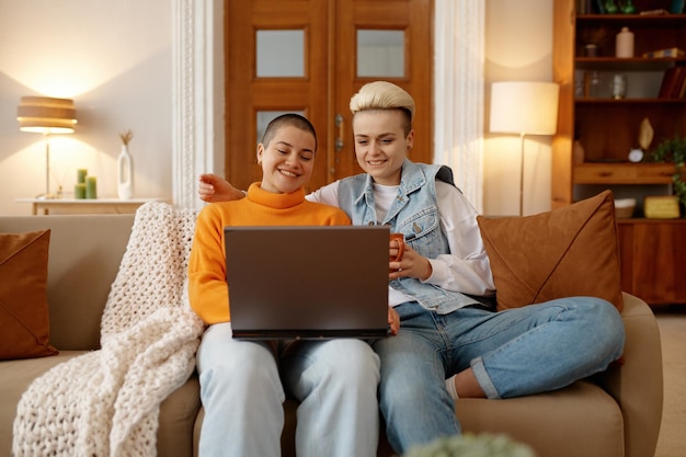 Happy loving lgbt woman couple watching video online using laptop while sitting and resting on sofa couch in cozy home living room