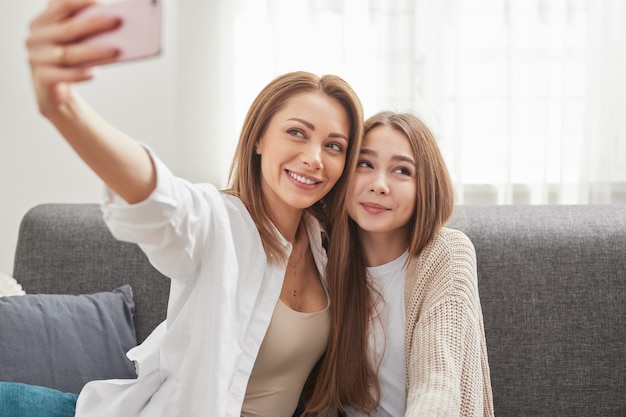 Happy loving mom with preteen daughter sitting on couch and taking selfie on smartphone while enjoying free time together at home