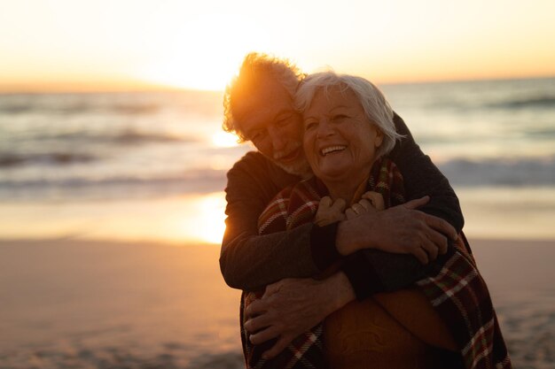 Happy, loving senior Caucasian couple relaxing on an empty beach, embracing at sunset, the woman wrapped in a blanket. Couple in retirement with healthy outdoor active lifestyle