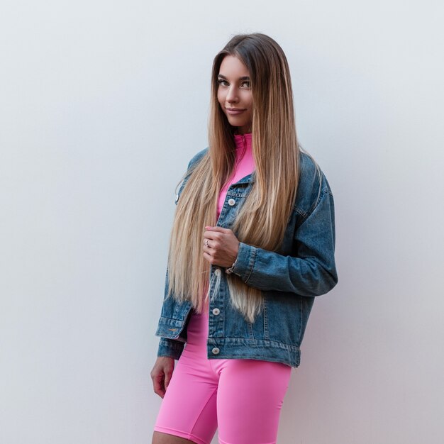 Happy luxurious young woman in a fashionable denim jacket in stylish pink shorts with beautiful smile poses outdoors near a vintage wall. Joyful urban girl outdoors on a warm summer day. Retro style.