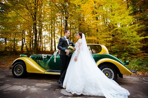 幸せな豪華な結婚式のカップルのキスをし、レトロな秋の花束の車でレトロに近い