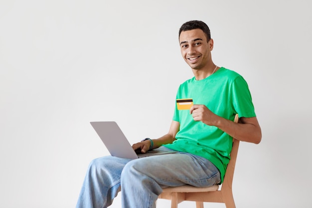 Happy male buyer using laptop and credit card making payment online sitting on chair over white wall free space