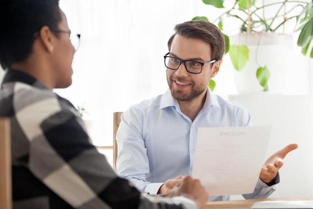 Happy male CEO or boss holding applicant resume talking with black female employee at interview HR manager satisfied with candidate experience African American girl make good first impression