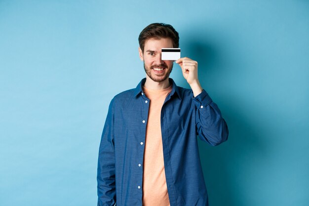 Happy male client holding plastic credit card on eye and smiling, standing in casual outfit on blue background.