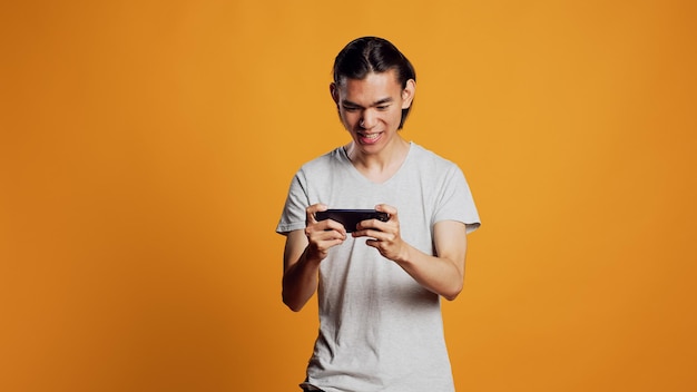 Happy male model playing video games on smartphone, using online gaming competition to have fun in studio. Young asian guy enjoying mobile game on phone, shooter challenge play.