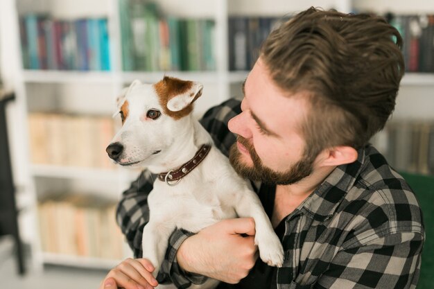 Photo happy male owner of jack russell terrier dog, feels responsibility of caring about pet, standing against bookshelves background. people and relationship with animals