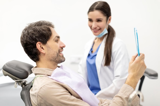 happy male patient looking in the mirror and checking teeth after professional dental treatment in